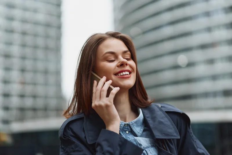 Happy young woman talking on smartphone outdoors in the city, smiling with eyes closed, wearing casual jacket and denim shirt. Concept of communication and lifestyle.