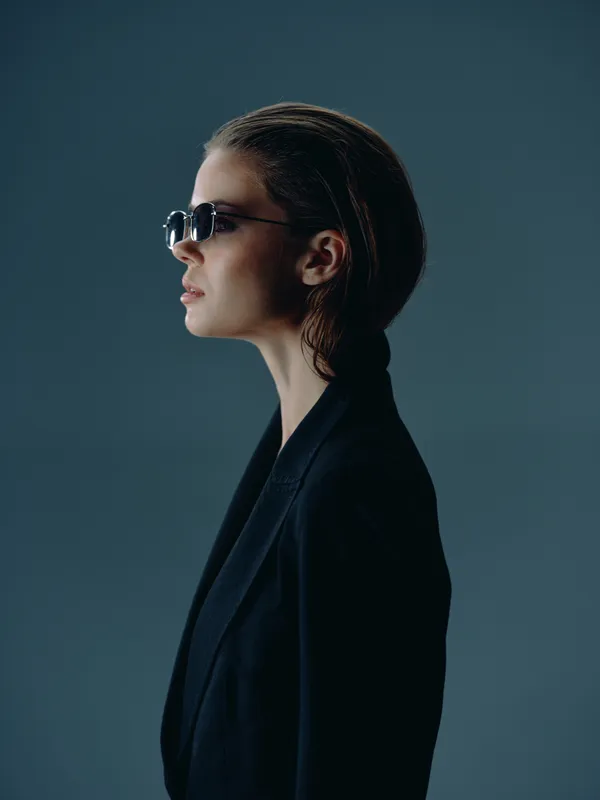 Stylish woman wearing sunglasses and a dark blazer, looking confident with a modern hairstyle against a muted blue background.