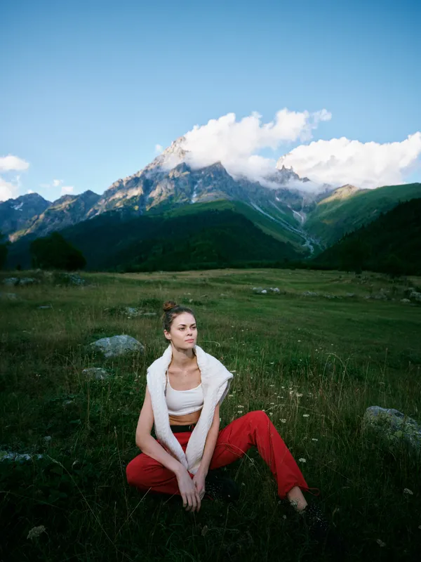 Meadow and mountains backdrop with expansive nature and clear sky, featuring a relaxed woman in casual wear sitting on grass, capturing a scenic outdoor moment of calm and style