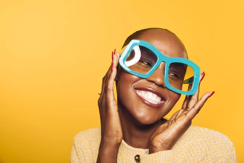 Happy African woman with dark skin wearing oversized blue glasses and yellow sweater smiles joyfully on bright yellow background. Concept of fashion, beauty, and positive mood.