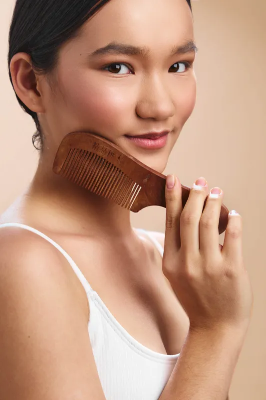 asian woman smiles softly as she styles her hair with a wooden comb, posed in a neutral studio, conveying calm elegance, personal care, and simple lifestyle beauty in a clear, intimate moment.