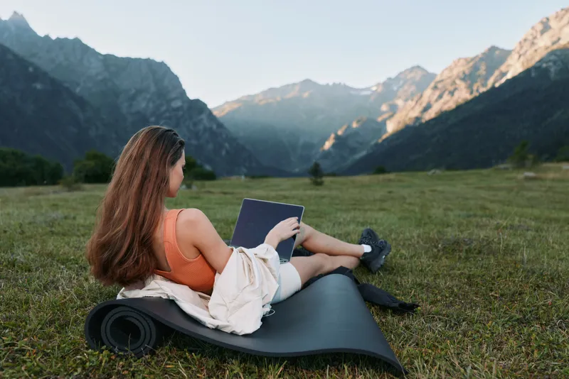 A relaxed woman lies on a yoga mat in a scenic meadow, working on a tablet. She wears casual clothes and enjoys peaceful, natural surroundings, creating a calm outdoor work vibe in bright daylight.