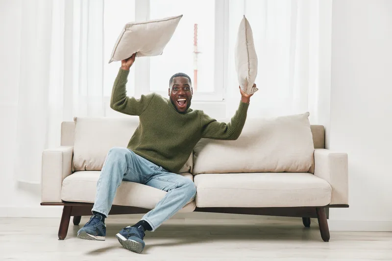 Happy man celebrating at home with pillows, wearing casual green sweater and jeans, joyful expression, modern interior, light colors, cozy atmosphere