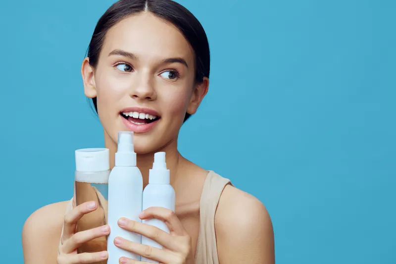 Young woman with a joyful expression holding skincare products against a blue background, promoting healthy skincare routines and beauty concepts