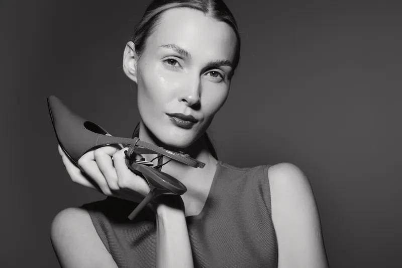 Portrait of a young Caucasian woman with fair skin holding high heel shoes near her face on dark background. Concept for fashion, elegance, and beauty.