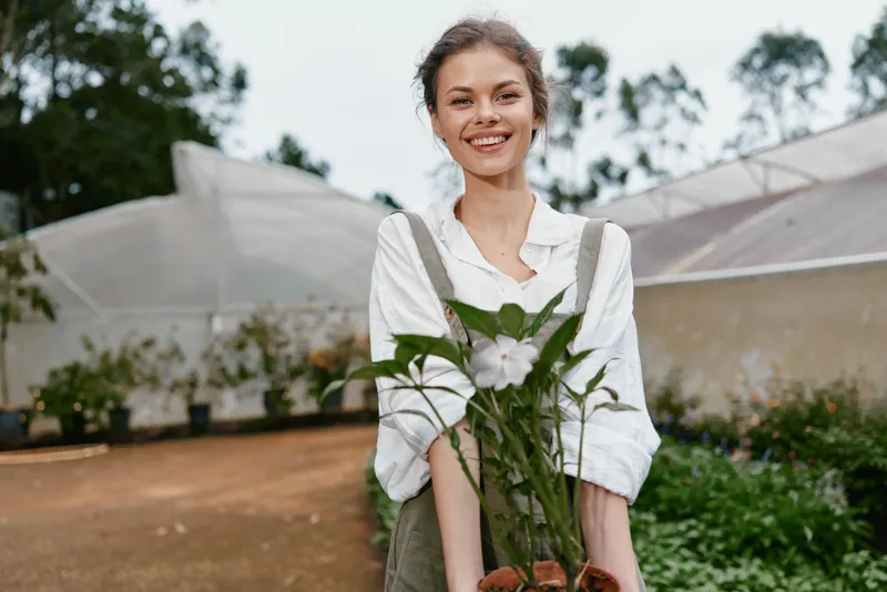 Smiling young woman holding potted green plants in a greenhouse, wearing casual work attire, showcasing a connection with nature and gardening