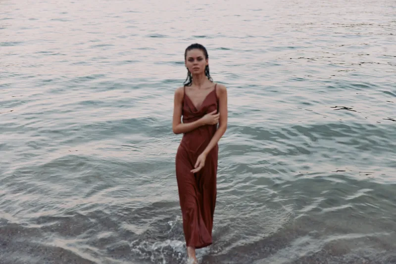 A woman in a deep red dress stands in shallow water at the shoreline, creating a bold focal point against the calm sea and soft, frothy waves under pale light.