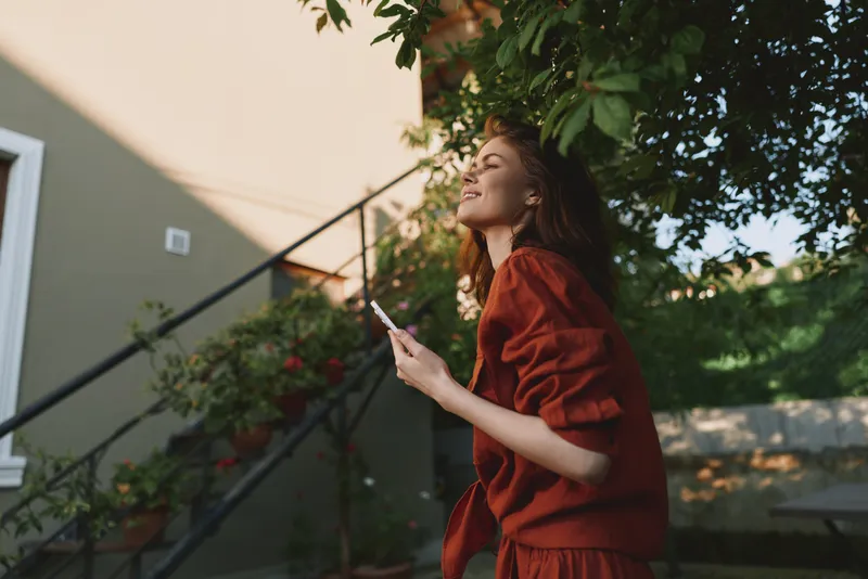 Woman lifestyle scene outdoors, casual red outfit, smiling while looking up and holding a phone, sunlit garden background, relaxed confident moment