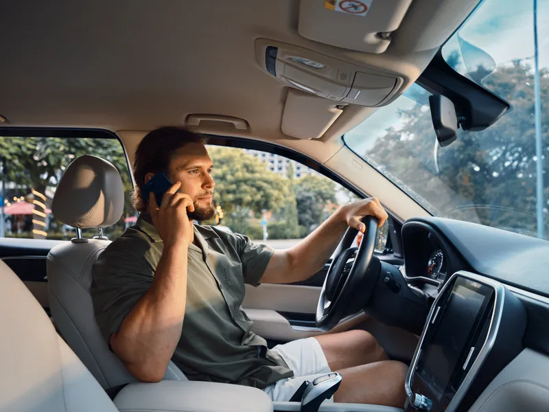 A focused male driver sits behind the steering wheel and talks on a smartphone, capturing a candid moment inside a modern car during daytime city drive.