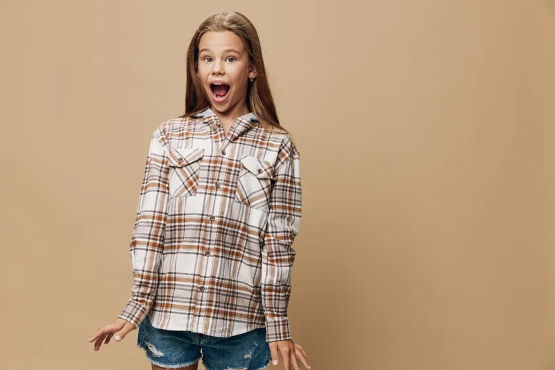 Girl standing with surprise expression wearing a plaid shirt and denim shorts on beige background. Child with open mouth showing excitement and casual clothes for childhood style.