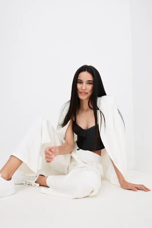 young woman sitting relaxed in white pants and blazer with black top, long straight dark hair, casual style, minimalistic white background, portrait fashion.