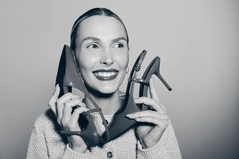 Smiling white woman with fair skin holds two high heel shoes near face, showing joy and fashion interest. Studio portrait with neutral background. Concept of style and happiness.
