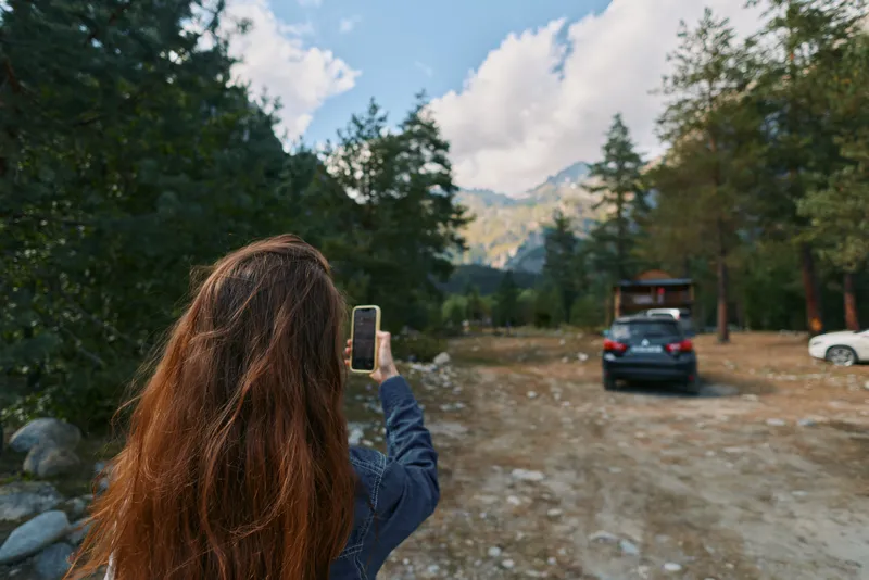 A woman with long hair stands outdoors, capturing a landscape with a smartphone. She faces mountains and trees, creating a calm and immersive nature scene.