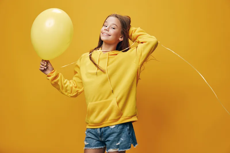 Girl with balloon smiling and posing in casual yellow hoodie and denim shorts, standing against orange background. Child playing, leisure, happiness, and fun in studio shot.