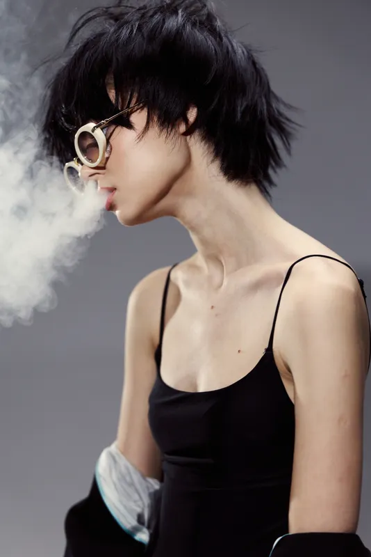 Young woman in sunglasses blowing smoke, wearing black dress with short hair and stylish edgy look