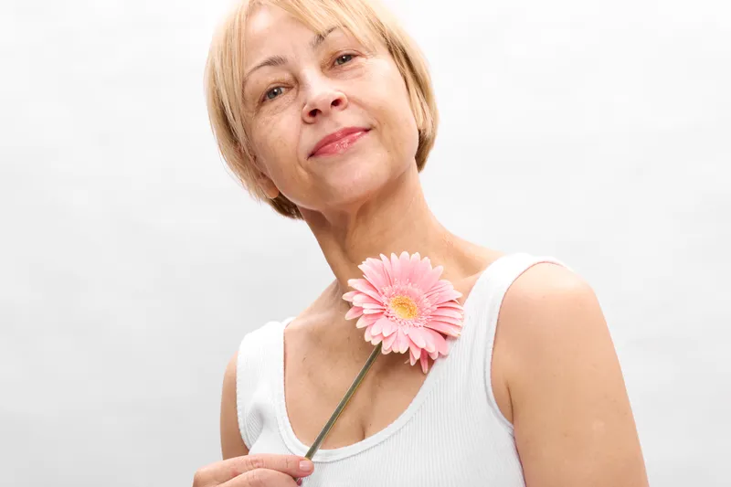 Elderly woman portrait holding a pink gerbera flower, white studio background, soft smile, calm spring mood, gentle light, closeup of face and bloom, peaceful nature theme