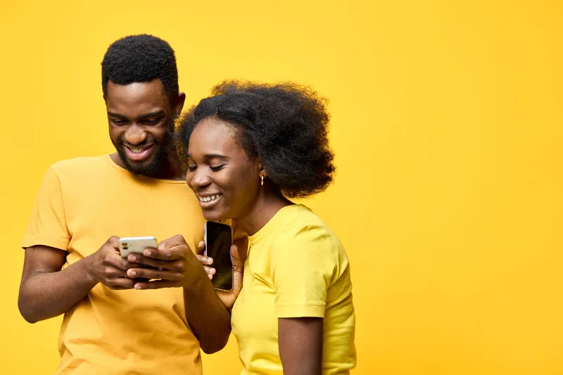 A cheerful young African couple sharing a joyful moment while using their smartphones against a vibrant yellow background, showcasing happiness and connection