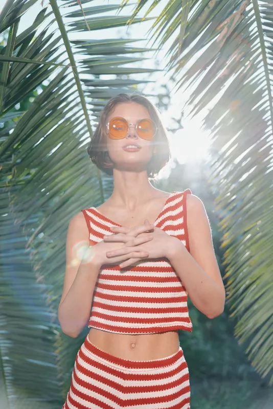 A stylish woman stands outdoors among palm leaves, wearing a red and white striped crop top and matching skirt. She wears round sunglasses and exudes confidence under bright tropical light.