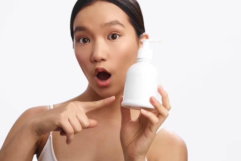 Asian woman holds a white pump bottle and points at it with surprised expression, presenting a skincare product in a clean studio setting and bright, neutral background.