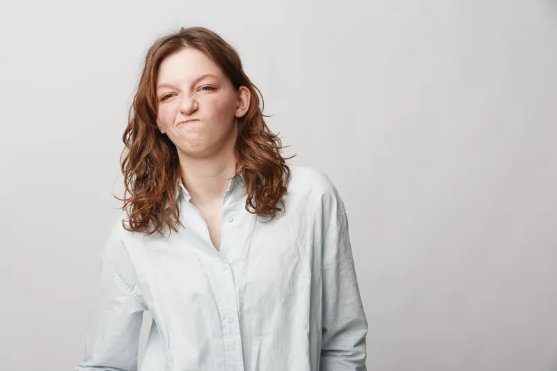 Young woman with curly hair wearing white shirt makes funny face with scrunched nose and pursed lips. Isolated on light background. Concept of humor, expression, and casual style.