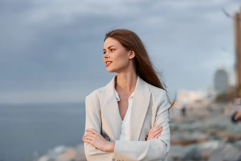 Confident woman in a business suit standing by the seaside, with a calm expression and natural wind-blown hair, embracing her entrepreneurial spirit.