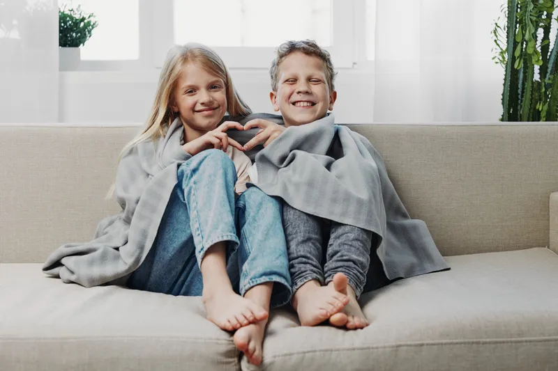 Two happy children wrapped in gray blanket sitting barefoot on sofa smiling and making heart gesture with hands in cozy bright living room.