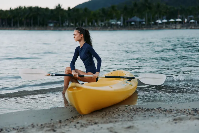 Young woman rowing a yellow kayak on the beach with calm sea and tropical background during the day.