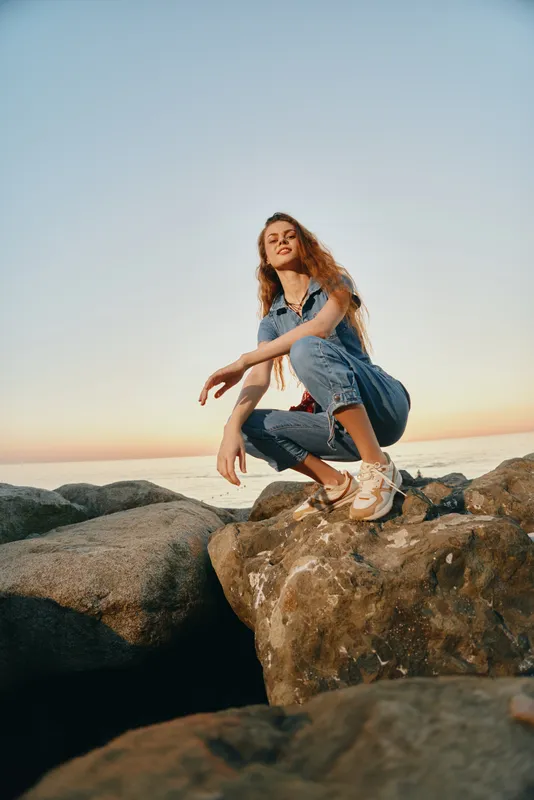 lifestyle woman in modern boho-western denim street style crouching on rocks by the sea during warm golden hour with film color effect and casual sneakers