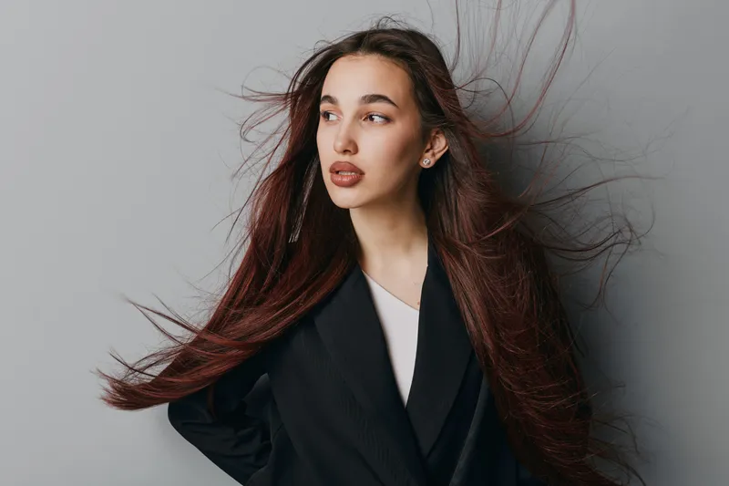 Young woman with long flowing hair and natural makeup posing in black blazer against gray background. Fashion portrait with confident expression and stylish casual outfit for beauty concept.