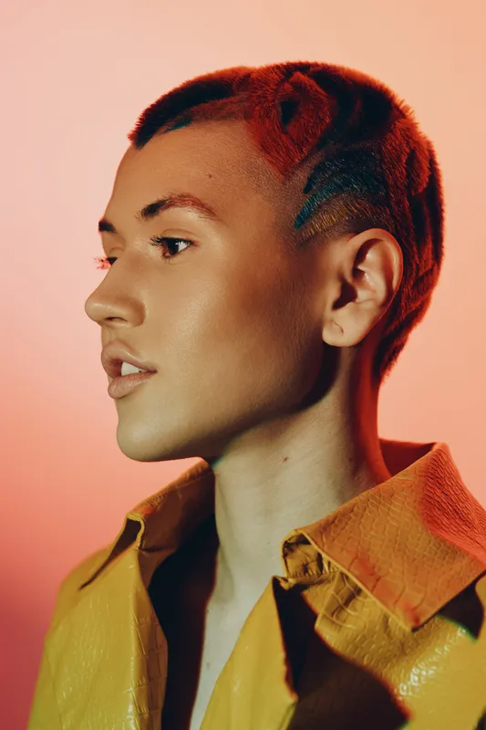 Close-up portrait of a young woman with a short, colorful haircut wearing a vibrant yellow shirt against a warm, peach background, showcasing bold fashion and style.