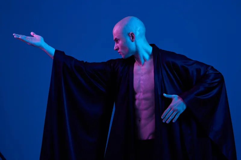 Artistic male figure with striking pose, wearing a flowing black robe. The scene is illuminated with blue lighting, creating a dramatic and captivating atmosphere.