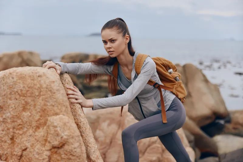 A focused young woman climbing rocky terrain by the sea, showcasing determination and adventure with a backpack, perfect for travel or fitness themes