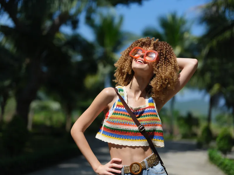 Smiling woman with curly hair wearing colorful crop top and oversized sunglasses outdoors in sunny tropical setting enjoying summer day.