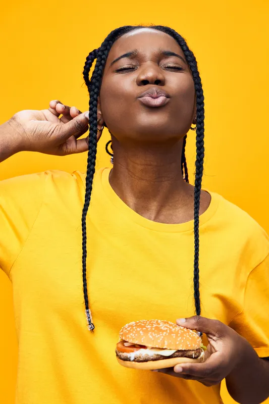 Young African woman enjoying a delicious burger against a vibrant yellow background, showcasing joy and enthusiasm for healthy eating and flavorful meals