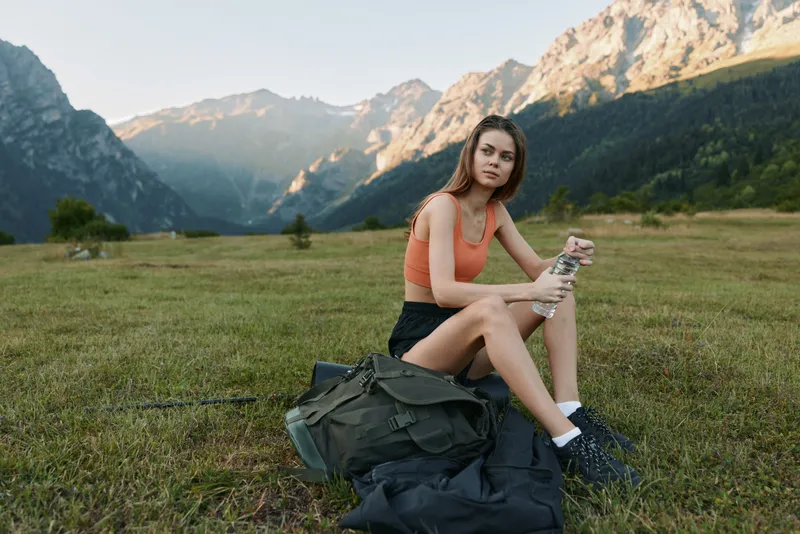 A young woman sits in a grassy meadow with rugged mountains in the distance, taking a break from hiking. She wears an orange top and dark shorts, looking relaxed and focused.