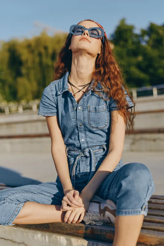 Lifestyle woman in modern boho-western denim street style wearing sunglasses, sitting outdoors with relaxed posture and warm film color tones in sunny urban environment.