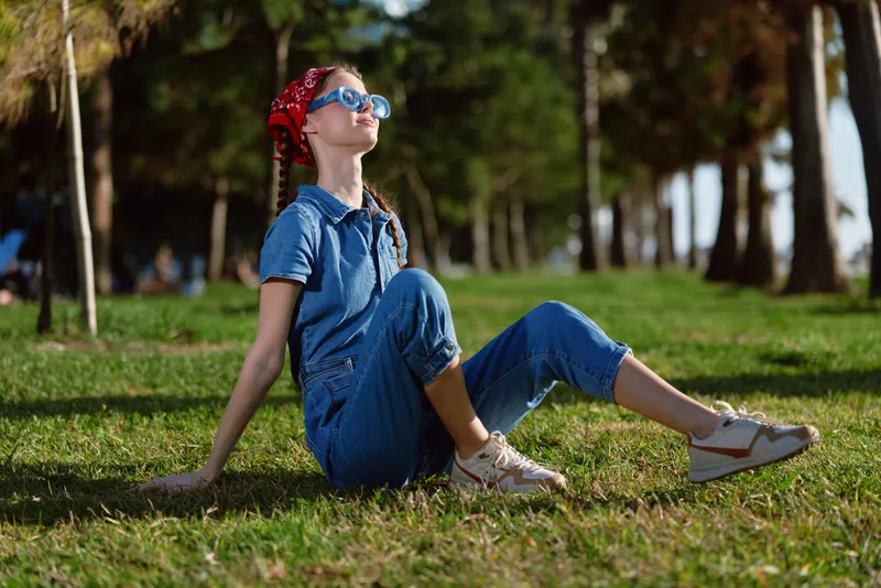 Lifestyle woman in modern boho-western denim street style wearing sunglasses and red bandana relaxing outdoors on grass in warm sunny day with film color effect.