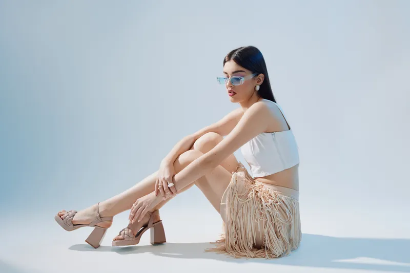 Young woman in stylish white top and fringed skirt sits on the floor wearing sunglasses and high heel sandals. Studio shot on light background. Concept of woman beauty fashion.