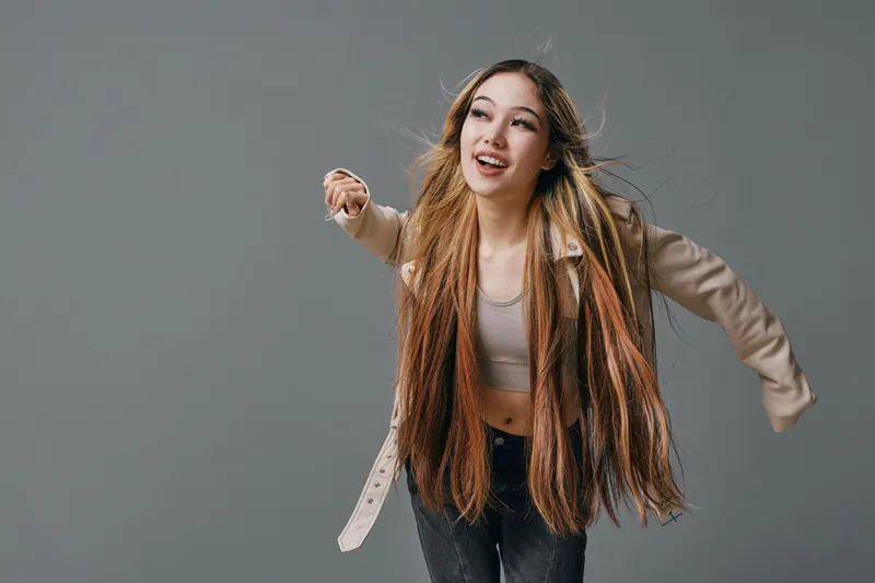 Young woman with long colorful braids dancing and smiling in casual clothes against gray background with joyful expression and dynamic pose.