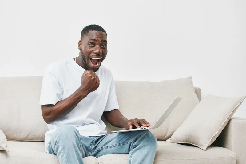 Excited young African man working on a laptop at home, showcasing joy and triumph with a neutral background and casual attire