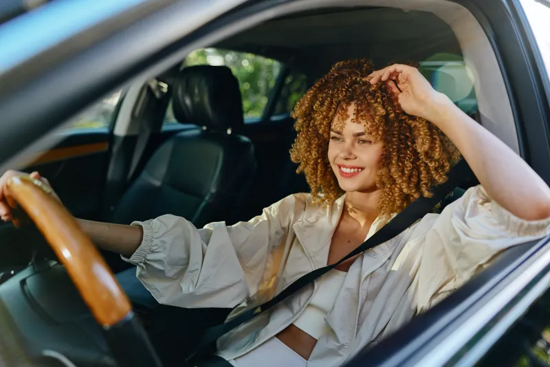 Smiling woman driving car with curly hair, wearing white clothes and seatbelt, enjoying sunny day and relaxed atmosphere inside modern vehicle.