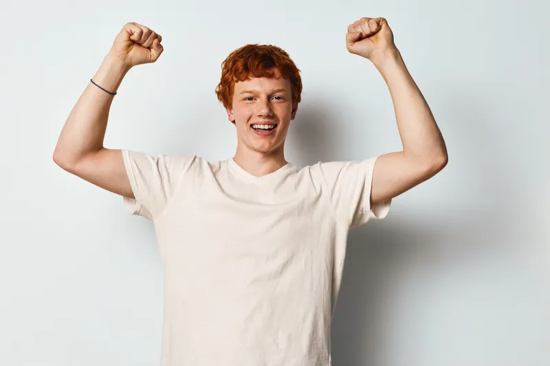 Young man with red hair and casual white t shirt raising fists in celebration for success and happiness on plain background. Portrait, smiling and positive emotion.