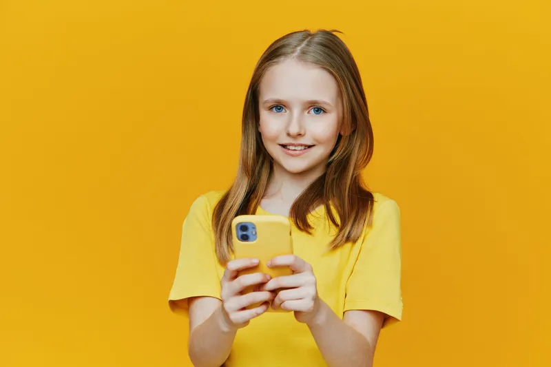 Cheerful girl smiling while holding a smartphone against a vibrant yellow background, showcasing a playful and positive mood in contemporary fashion