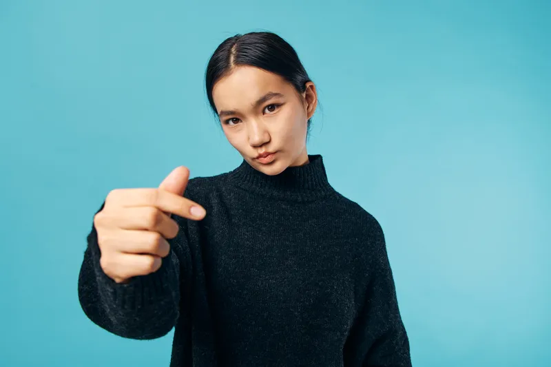Young woman with dark hair wearing black sweater pointing finger forward against blue background. Female person with serious expression and casual clothing in studio portrait.