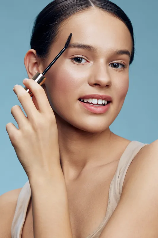 Young woman applying mascara with a smile, showcasing beauty, confidence, and skincare in a soft blue background, highlighting makeup and self care trends