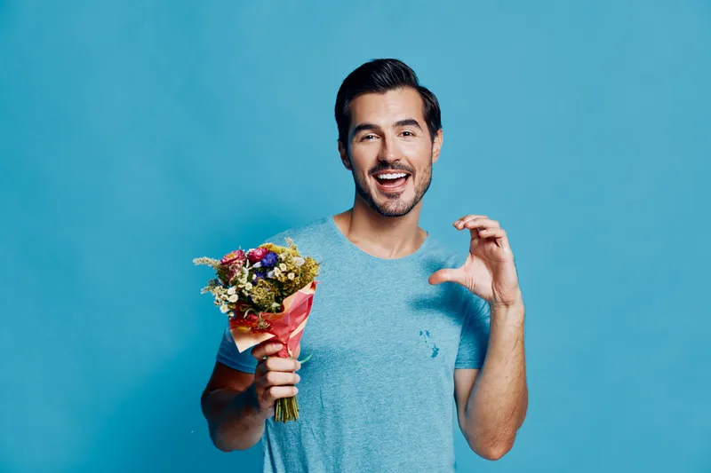 Cheerful smiling man holding a bouquet of flowers while looking at the camera, dressed in a casual blue t-shirt, isolated on bright blue background. Lifestyle concept