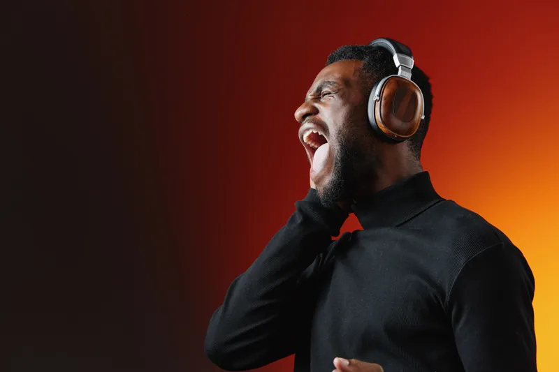 Young Black man singing passionately with headphones, against a vibrant gradient background showcasing orange and dark tones, evoking emotions of joy and energy