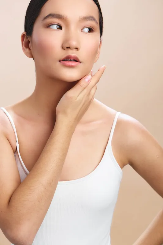 Asian woman with a thoughtful pose wearing a white tank top, captured in a neutral studio setting. The soft lighting highlights her skincare and calm expression, conveying poise, confidence, and a