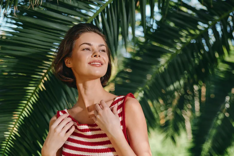 A cheerful woman poses outdoors among palm trees, wearing a striped tank top. She looks relaxed and confident, enjoying a sunny moment in a tropical setting.