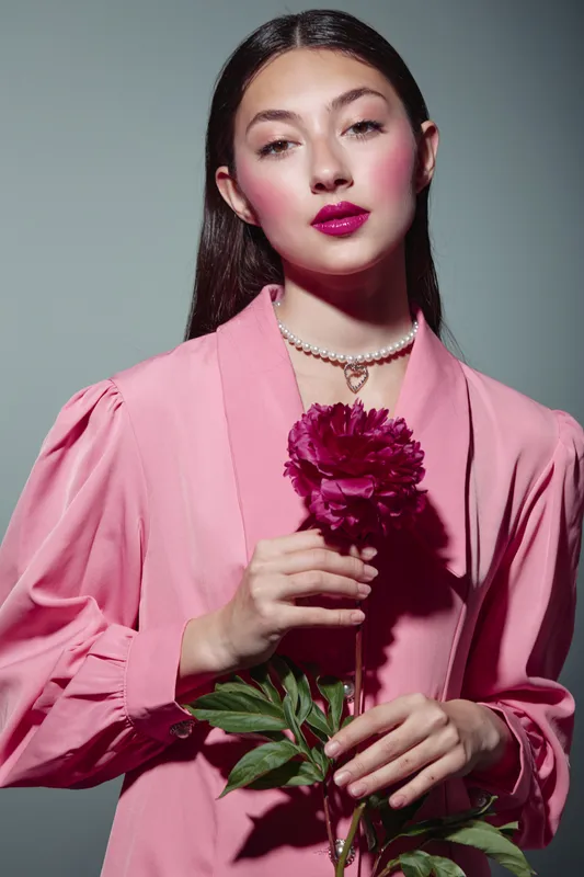 Young woman in pink blouse holds dark pink flower posing confidently against gray background. Concept of beauty, fashion, creative style, and floral elegance.